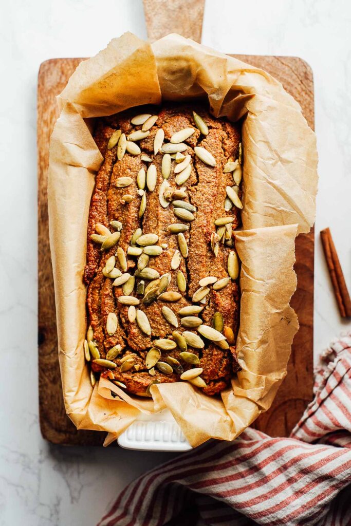 Cooked bread sprinkled with pumpkin seeds in a loaf pan lined with brown parchment paper.