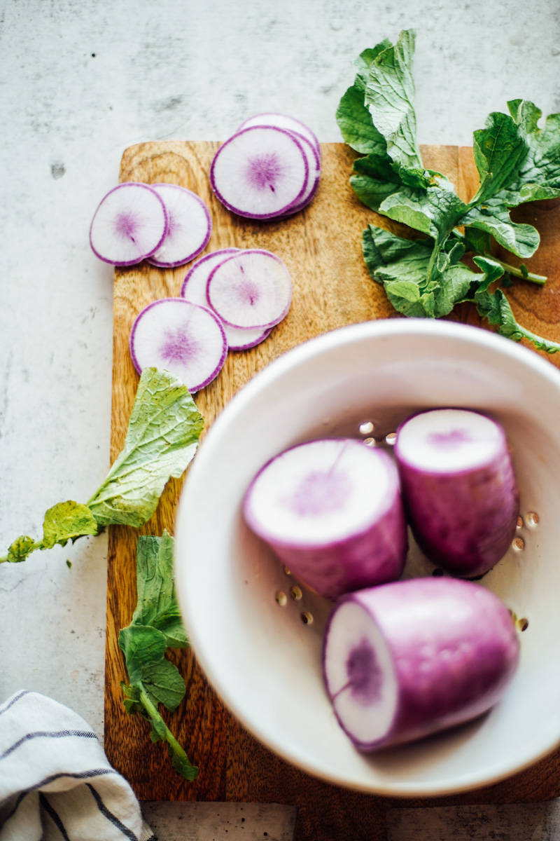 Purple watermelon radishes cut in half on a cutting board with radish tops on the board.