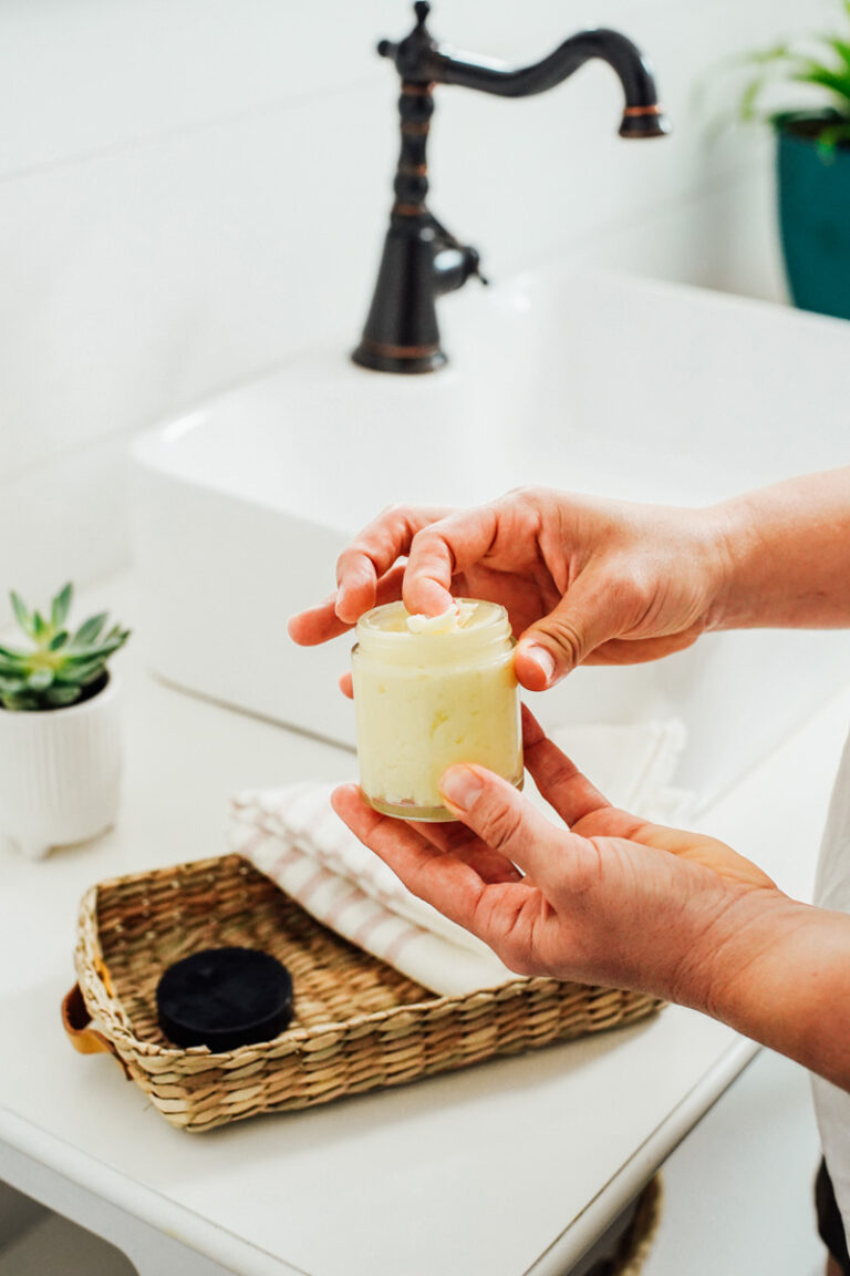 Scooping shea butter lotion from a clear glass jar using pointer finger.