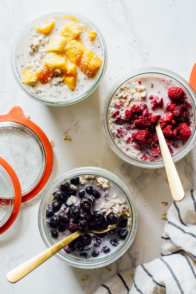 Three jars of overnight oats on the counter with frozen fruits on top: mango, raspberries, and blueberries.