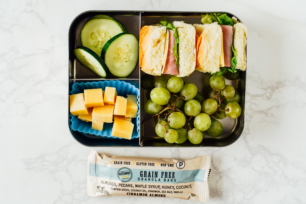 School lunch: ham sandwich on a bun with cheese cubes, grapes, and cucumbers.