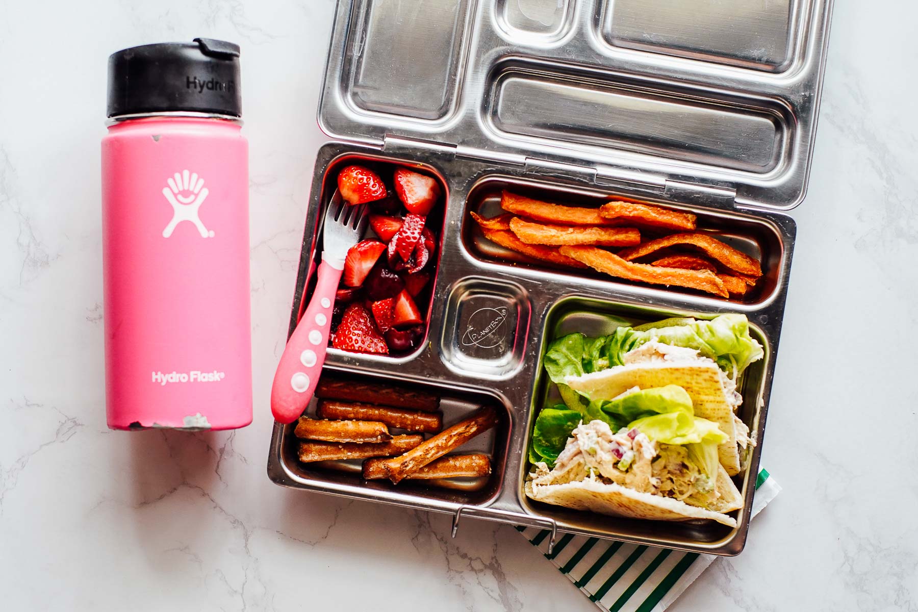 Chicken salad in pita bread with pretzels, strawberries, and carrot fries in a lunchbox.