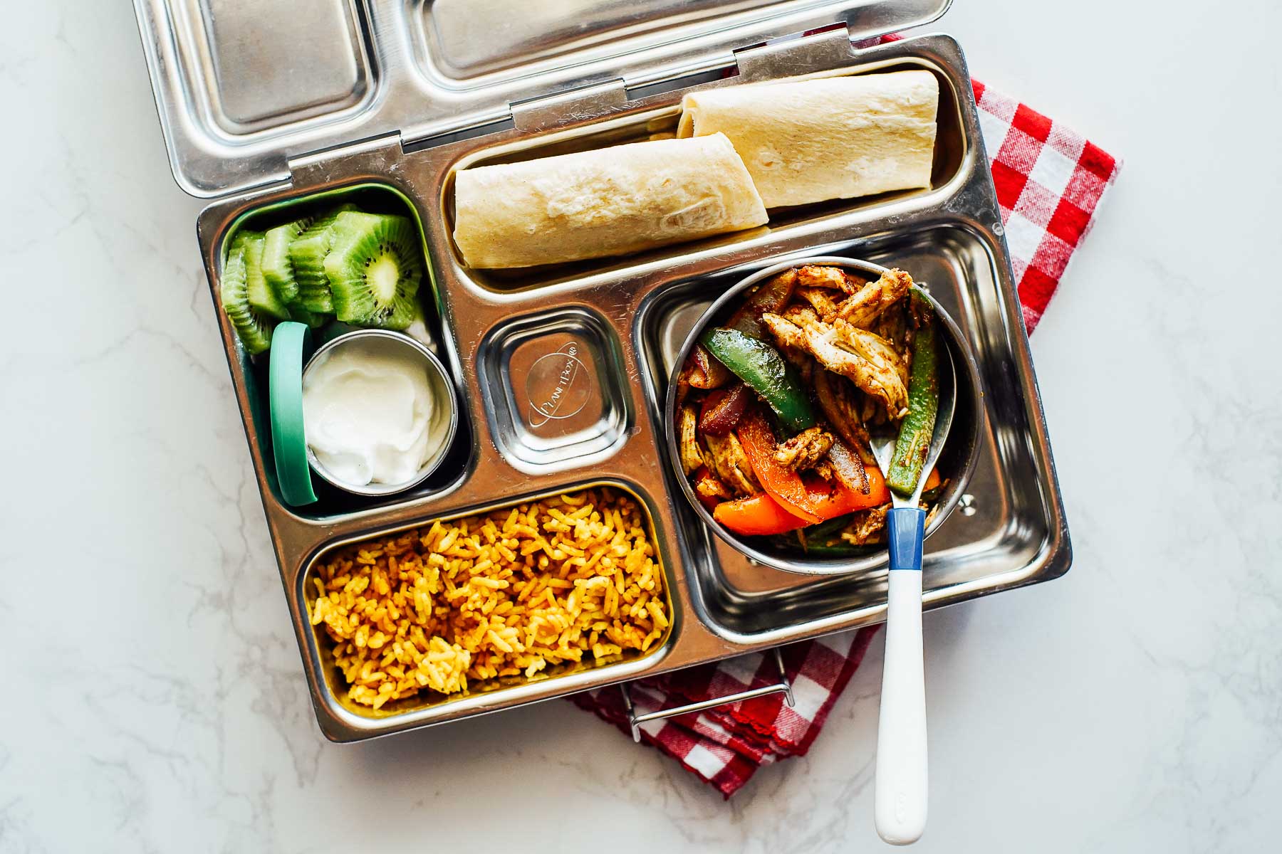 Leftover chicken fajitas with yellow rice, a tortilla, and yogurt with kiwi slices in a lunchbox.