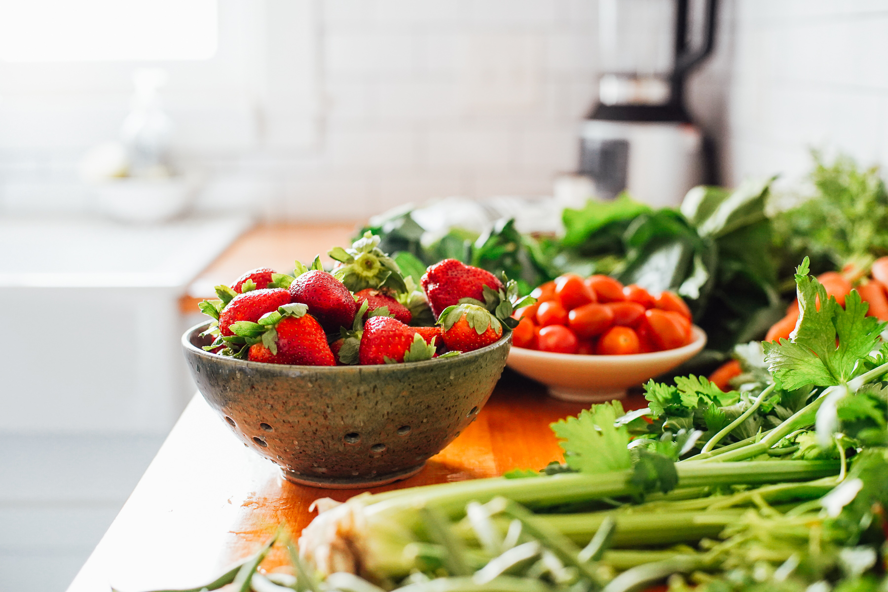 Strawberries in a colander on the kitchen counter with other veggies.